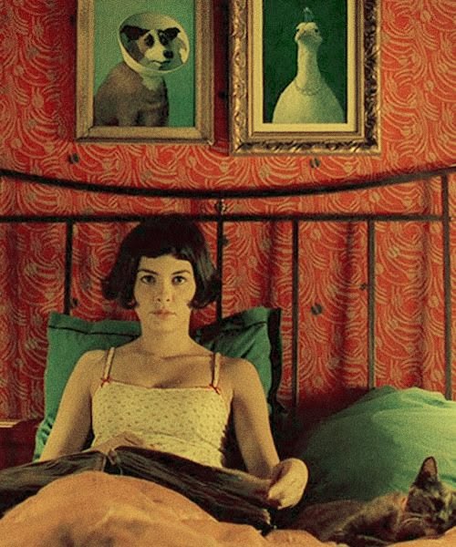 Amélie sits centred on her bed, framed by red patterned walls, with green pillows around her.