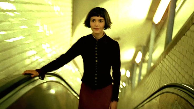 Amélie rides an escalator through a yellow-green metro passage, the rails and lights leading into the frame.