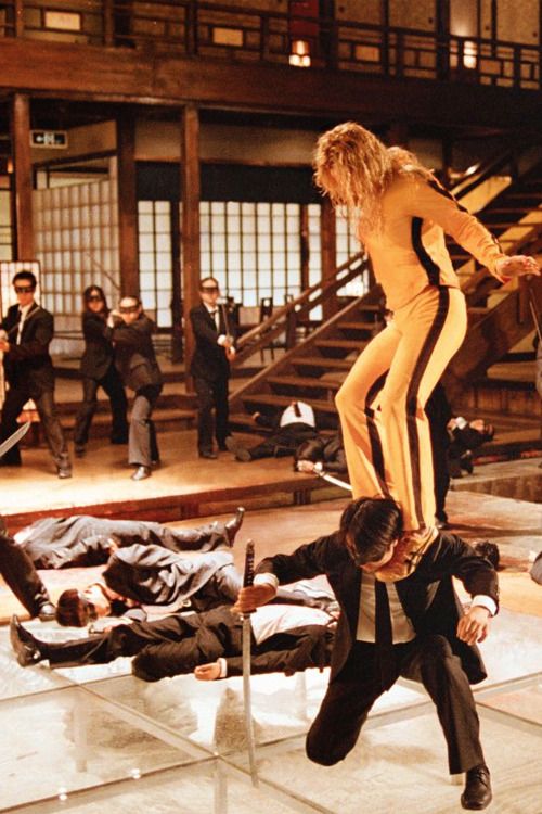 The Bride in her yellow suit stands among fallen fighters in the warm-lit House of Blue Leaves hall.